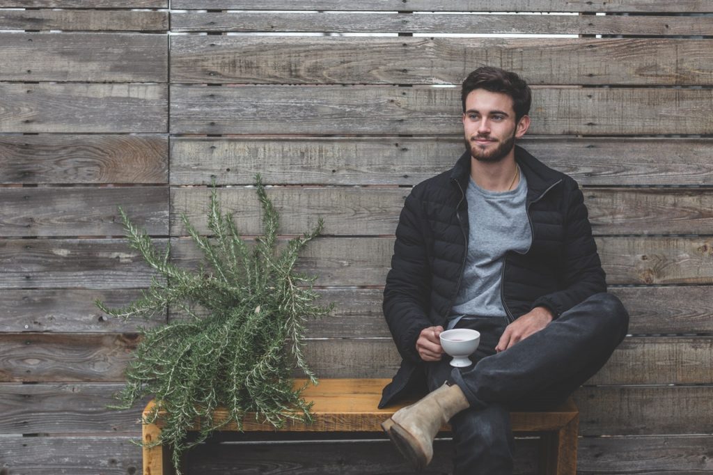 homme brun portant un t shirt gris sous une doudoune foncée un jean et des boots claires avec une tasse à la main